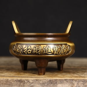 May include: A bronze-toned metal incense burner featuring three short legs and two upright handles. The exterior body displays a central rectangular panel with embossed Chinese characters reading 招財進寶, flanked by decorative floral relief carvings.