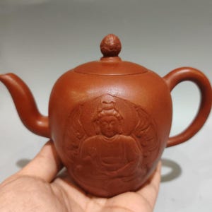 May include: A reddish-brown clay teapot featuring a relief carving of a multi-armed deity on the front. The teapot has a curved spout, a rounded handle, and a lid topped with a small, textured knob. The surface has a matte, earthy finish.