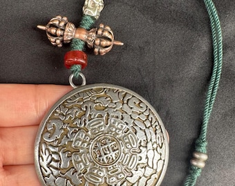 Puede incluir: Colgante plateado con un diseño circular intrincado. Está unido a un cordón verde con un elemento decorativo y una pequeña cuenta roja. El diseño del colgante presenta un patrón central rodeado por un borde ornamentado.