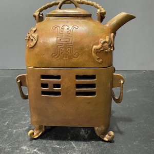 Puede incluir: Una tetera de latón con un asa decorativa y una base de quemador. La tetera tiene caracteres chinos y un diseño de dragón.