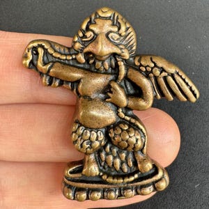 May include: A bronze figurine of a mythical creature with a detailed face, wings, and ornate clothing. The figure is posed with arms outstretched, holding a snake-like object. The base of the figurine has a decorative pattern.