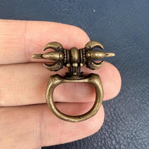 Pode incluir: Um pequeno puxador de gaveta de cor latão antigo com uma pega em anel. O puxador decorativo apresenta um design central com elementos de três pontas em cada lado. O design geral é ornamentado e detalhado.
