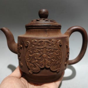 May include: Brown ceramic teapot with a lid and handle. The teapot features carved dragon designs on the front and sides. The lid has a small knob and decorative cutouts. The spout is curved, and the handle is a rounded loop.