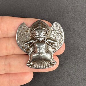 May include: A silver metal pendant depicting a mythical creature with wings, a bird-like head, and a human-like body. The pendant is held in a hand against a dark background. The design is detailed and appears to be vintage, with intricate detailing.