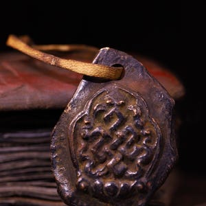 May include: A weathered, bronze-colored pendant with an intricate, embossed design and a leather strap. The pendant is rectangular with rounded edges and a loop for the strap. The background shows a stack of aged, red-toned pages.
