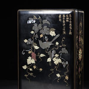 May include: A rectangular black lacquer box featuring intricate mother-of-pearl and gold inlay depicting a bird perched on a grapevine. The surface displays detailed leaf patterns, clusters of grapes, and elegant Chinese calligraphy characters.