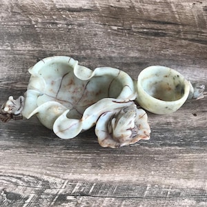 May include: A carved jade inkstone with a small round well and a larger, leaf-shaped well. The inkstone is a light green color with brown and white streaks.