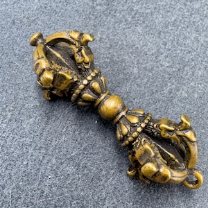 Peut inclure: Un vajra en laiton de style antique, un objet rituel avec des détails ornés. Le vajra présente une sphère centrale, des motifs complexes et des figures stylisées en forme de dragon à chaque extrémité. L'objet est posé sur une surface grise.