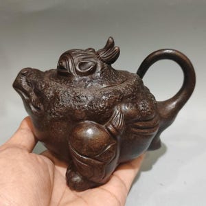 May include: A dark brown clay teapot shaped like a mythical creature with a textured body, a curved handle, and a lid featuring sculpted details. The teapot has a spout on the left side and a rounded base, showcasing a matte finish and intricate carvings.