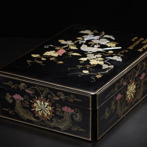 May include: A rectangular black lacquer box featuring an inlaid mother-of-pearl design on the lid depicting a bird perched among grapevines. The sides of the box are decorated with painted floral motifs in gold, pink, and white, with Chinese calligraphy.