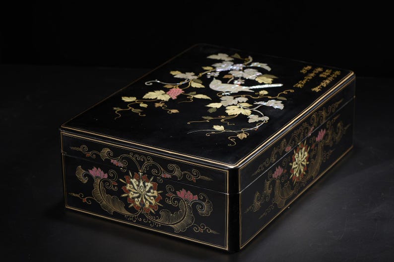 May include: A rectangular black lacquer box featuring an inlaid mother-of-pearl design on the lid depicting a bird perched among grapevines. The sides of the box are decorated with painted floral motifs in gold, pink, and white, with Chinese calligraphy.
