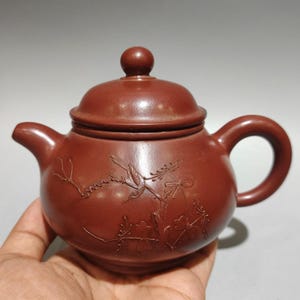 May include: A reddish-brown clay teapot featuring a rounded body, a matching lid with a small knob, and a curved handle. The exterior surface is decorated with an engraved design of a bird perched on a flowering branch with delicate blossoms.