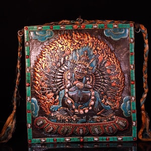May include: A detailed, rectangular, ornate religious artifact with a central figure surrounded by flames. The figure has multiple arms and is seated. The frame is green with turquoise and red accents. Braided cords hang from the top corners.