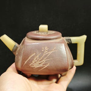 Pode incluir: Uma pequena chaleira marrom com uma alça, bico e tampa de cor clara. A chaleira tem uma forma geométrica e um design decorativo gravado na frente. A tampa tem um pequeno botão quadrado. A chaleira é provavelmente feita de cerâmica ou pedra.