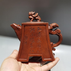May include: A reddish-brown, intricately carved teapot with a unique handle and spout design. The teapot features detailed carvings, including a small animal figure on the lid and Chinese characters on the body. The teapot is held in a hand.