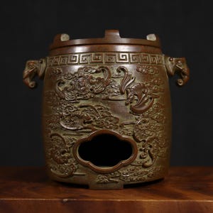 Puede incluir: Un quemador de incienso de bronce con intrincados tallados de dragones y nubes. El quemador tiene una parte superior cuadrada con un dise&ntilde;o de llave griega y dos asas en forma de drag&oacute;n.