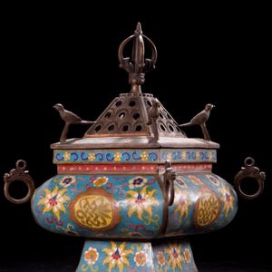 Könnte beinhalten: Ein verzierter, antiker Räuchergefäß mit einem türkisfarbenen, roten und gelben Blumenmuster. Die Metallarbeiten weisen zwei Vogelfiguren, eine dekorative Oberseite und Ringgriffe auf. Der Sockel ist quadratisch, der Deckel pyramidenförmig.