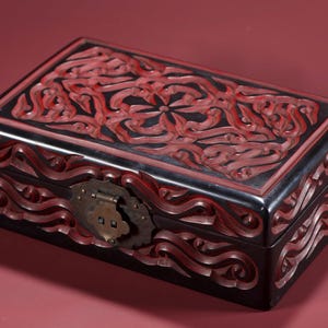 Puede incluir: Una caja negra rectangular con intrincados diseños rojos tallados en la tapa y los lados. La caja presenta un cierre metálico decorativo. La estética general sugiere un estilo vintage o antiguo, posiblemente para joyas o recuerdos.