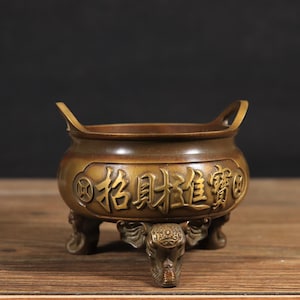 May include: A bronze-colored incense burner with handles and elephant-shaped feet. The burner has Chinese characters engraved around the body. The background is a dark, solid color.