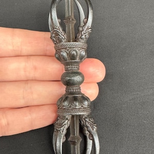 May include: A detailed, dark metal vajra, a ritual object with ornate detailing. The vajra features a central shaft, rounded midsection, and claw-like prongs at each end. The object is held against a dark background.