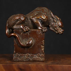 May include: A detailed bronze sculpture of a mythical creature, possibly a dragon, perched atop a rectangular base. The sculpture features intricate scales, horns, and a flowing tail. The base has a wave pattern at the bottom.