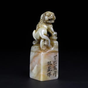 Op de afbeelding: Een uit steen gesneden zegel met een gehurkte leeuwfiguur bovenop een rechthoekige basis. De steen vertoont gemarmerde crème, beige en bruine tinten. Zwarte Chinese karakters zijn verticaal gegraveerd op de voorzijde van het gepolijste blok.