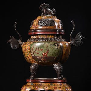 Puede incluir: Un quemador de incienso antiguo y ornamentado con tapa, que presenta una escena detallada de un paisaje con un ciervo. El quemador está sostenido por un soporte decorativo. La paleta de colores incluye dorado, rojo, verde y marrón, con intrincados patrones y asas con forma de animal.