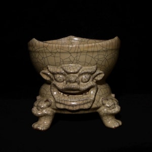 Może przedstawiać: Kremowa ceramiczna miska z krakelą, wspierana przez uśmiechnięte mityczne stworzenie z czterema nogami. Misa ma rozszerzoną krawędź i jest umieszczona na czarnym tle.