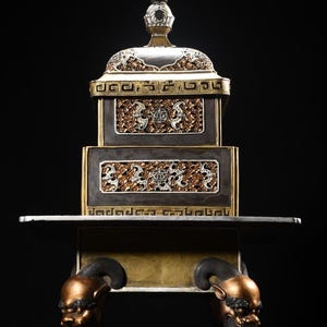 Op de afbeelding: Een sierlijk, meerlagig decoratief object in zilver en goud. De bovenkant heeft een gewelfd deksel met gedetailleerde gravures. De basis heeft twee niveaus met ingewikkelde reliëfpatronen, ondersteund door poten met bronzen, gestileerde dierenkoppen.