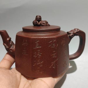 May include: A brown ceramic teapot with a lid and handle. The teapot has a spout and is decorated with carved Chinese characters. A small animal figure sits atop the lid.