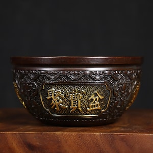 May include: A dark brown bronze bowl with intricate carvings and gold accents. The bowl has a raised rim and a circular base. The inscription "東 鎮 金" is visible on the front of the bowl.