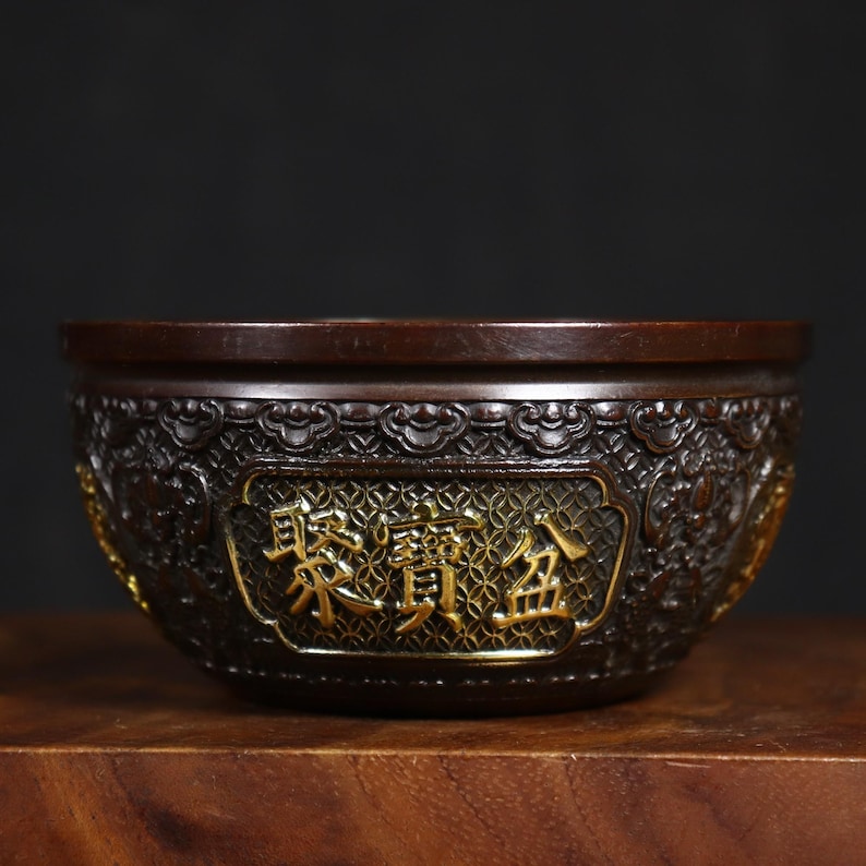 May include: A dark brown bronze bowl with intricate carvings and gold accents. The bowl has a raised rim and a circular base. The inscription "東 鎮 金" is visible on the front of the bowl.