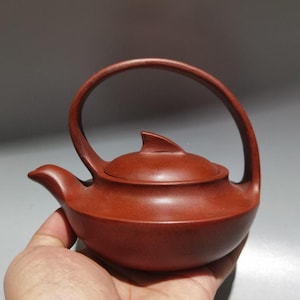 Puede incluir: Una pequeña tetera de cerámica de color marrón rojizo con un asa curva y una tapa. La tetera tiene un pico y un detalle en forma de aleta en la tapa. La tetera se sostiene en una mano.