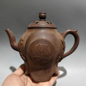 Può includere: Teiera in ceramica marrone con coperchio e manico. La teiera ha un corpo rotondo con un emblema circolare e caratteri cinesi. Il beccuccio e il manico sono curvi. La teiera è tenuta in una mano.