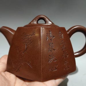 May include: A brown ceramic teapot with a geometric design. The teapot features a spout, handle, and a small loop on the lid. The body of the teapot has Chinese characters and a floral design etched into the surface.