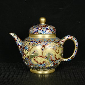 May include: A decorative teapot with a lid, featuring a gold dragon design on a multicolored background. The teapot has a gold handle, spout, and base. The lid is topped with a gold knob. The design includes blue, red, and green accents.