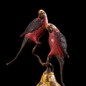 Puede incluir: Escultura decorativa de bronce de dos loros con cuerpos rojos y detalles negros, posados en una rama sobre una base amarilla en forma de calabaza. La base está adornada con patrones florales y de nubes.