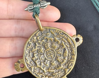 Puede incluir: Un colgante de color bronce con intrincados diseños en relieve, suspendido de un cordón verde. El colgante circular presenta un símbolo central rodeado de patrones detallados. El cordón está adornado con detalles metálicos plateados.