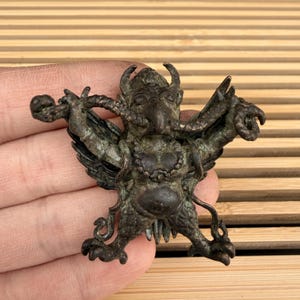 Op de afbeelding: Een bronzen beeldje van een mythisch wezen met een vogelachtig gezicht, hoorns en vleugels. Het figuur heeft uitgestrekte armen en houdt een klein voorwerp vast. De sculptuur heeft ingewikkelde details en een verweerd uiterlijk, wat wijst op een antiek of vintage stuk.