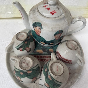 May include: A white porcelain tea set with a red and green design featuring two men shaking hands. The set includes a teapot, four cups, and a saucer. The text on the teapot reads "毛主席和他的亲密同志检阅文化革命大写" and the text on the saucer reads "安和他的亲密同志检阅文化革命大写".