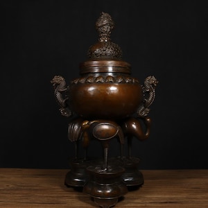 Peut inclure: Un brûle-encens en bronze avec un motif de dragon et une base à trois pieds. Le brûleur est décoré de détails complexes et possède un couvercle avec un fleuron.