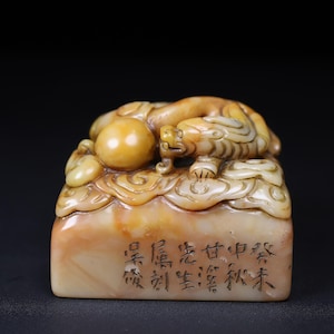 May include: A carved stone seal with a detailed tiger design on top. The seal is primarily beige and yellow with intricate carvings. The base of the seal has Chinese characters etched into it. The background is black.