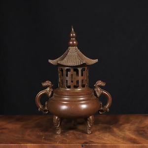 May include: A bronze-colored incense burner with a pagoda-style top and dragon-shaped handles. The burner has a round base supported by three feet. The top features a detailed, openwork design. The overall aesthetic suggests an Asian-inspired decorative piece.