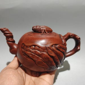 以下が含まれることがあります： 詳細な彫刻デザインが施された赤茶色のティーポット。 ティーポットは、ねじれた注ぎ口、湾曲したハンドル、上部に小さな昆虫のフィギュアが付いた蓋が特徴です。 ティーポットの本体は、彫刻された葉の模様で装飾されています。