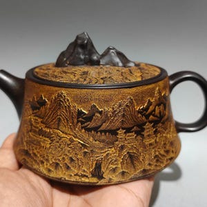 Puede incluir: Una tetera de cerámica marrón con un diseño detallado de paisaje montañoso tallado en el cuerpo. La tapa presenta un pico de montaña, y el asa y el pico son de color marrón oscuro.
