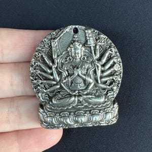 Puede incluir: Colgante plateado que representa una deidad sentada con múltiples brazos. El colgante tiene forma circular con intrincados grabados en relieve y un pequeño agujero en la parte superior para colgarlo. El diseño es complejo.