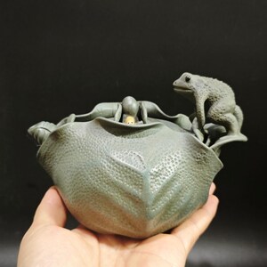 May include: A detailed, gray ceramic frog sculpture perched on a textured, leaf-shaped bowl. The bowl has a small figure inside. The sculpture is held in a hand against a dark background.