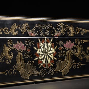 May include: A rectangular black box featuring a central floral motif in gold, red, and pink. The design includes intricate swirling gold line work and symmetrical leaf patterns that extend horizontally across the front surface of the box.