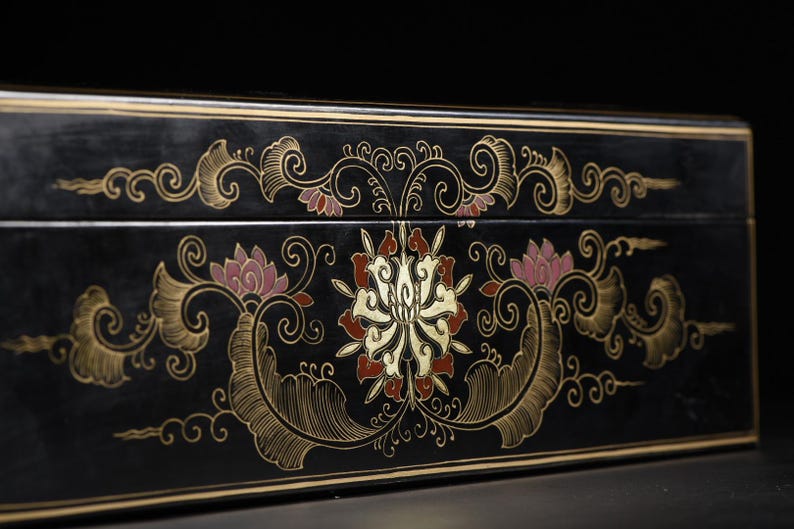 May include: A rectangular black box featuring a central floral motif in gold, red, and pink. The design includes intricate swirling gold line work and symmetrical leaf patterns that extend horizontally across the front surface of the box.