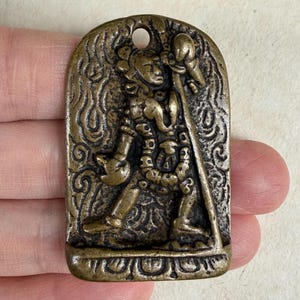 Puede incluir: Colgante de bronce con un relieve detallado de una figura que sostiene un bastón. La figura está adornada con intrincados patrones y se encuentra dentro de un marco arqueado. Un pequeño agujero en la parte superior para colgar.